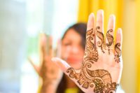 upclose view of a palm of a hand decorated with mehndi and wearing a wedding ring, with a blurry image of a woman in the background