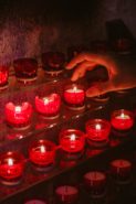 a hand reaching toward one votive candle on a shelf of lit candles in red glass containers
