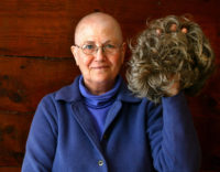 older white woman with glasses who's bald, holding a gray wavy haired wig in her hand
