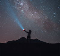 A night sky with a lot of stars. The dark silhouette of a person standing on a ridge extending a flashlight beam to the sky.