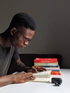 a man reading a book. the man is black with short hair and dark rimmed glasses. a stack of books sits on the table he's reading on.