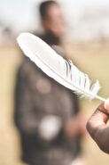 a hand holding a white feather out to a out of focus figure in the distance