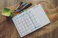 a planner lays open to the month of January, marked with a rainbow of sticky note flags. beside it lay a few markers and a green ceramic pilar with a star cut out of its side.