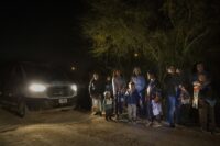 a group of 11 migrants, including six children, stand in the car lit only by the headlight of a white van parked to the left