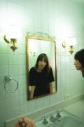 a woman in a black tshirt looks at the camera through her reflection in a gold trimmed bathroom mirror, the bathroom tile is mint green and the mirror is flanked by two globe lights