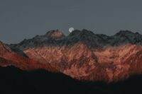 dusky mountains, with a run of red trees up the slope of the mountain furthest left. above them a full moon is almost completely risen.